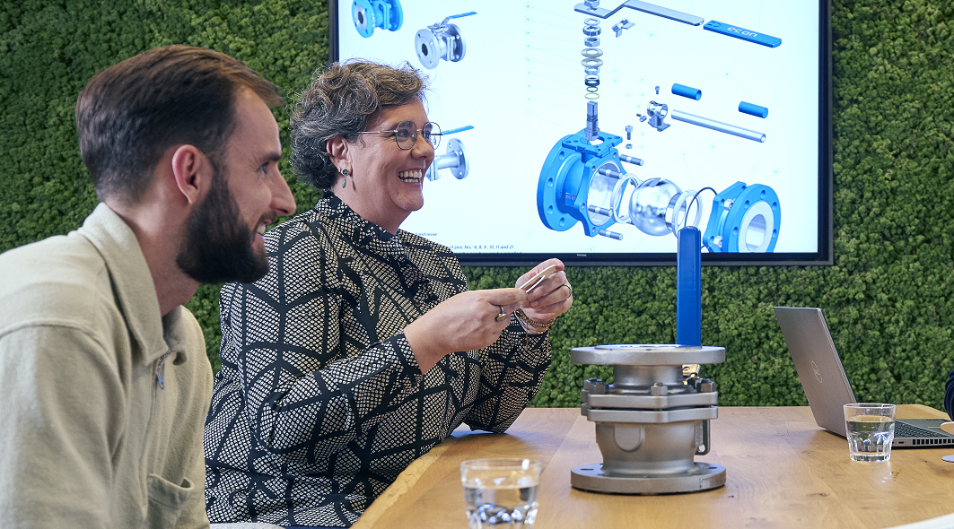 An image captures a cheerful moment where a man and a woman are seated at a large table, deeply engaged in a discussion about a mechanical product placed before them. Both individuals are smiling, indicating a positive and collaborative conversation. In the background, a television screen is visible, adding a modern, technological element to the setting. This scene highlights teamwork, innovation, and the exchange of ideas in a professional environment.
