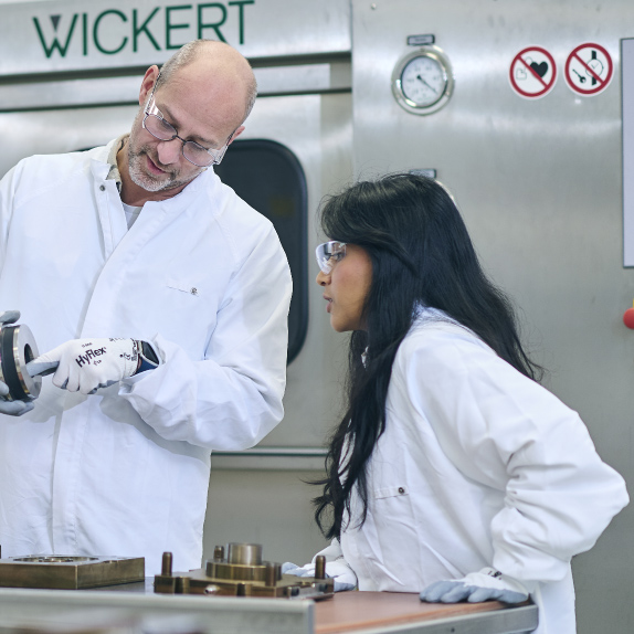 An image portrays two professionals, a man and a woman, both dressed in white laboratory coats and wearing protective glasses, deeply engaged in examining and discussing a metal component. They stand in a well-equipped laboratory, surrounded by various scientific instruments and equipment, emphasizing the precision and technical expertise required in their work. This setting reflects a collaborative research and development atmosphere, where innovation and meticulous attention to detail are paramount.