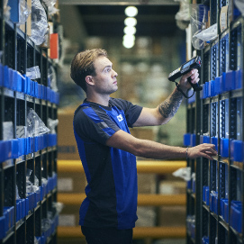 A young man stands in the center of a warehouse, intently working as he scans products with a digital scanner. He is surrounded by shelves filled with various items, highlighting the busy and organized environment of the warehouse. The focus on the young man and his task illustrates the important role of technology in inventory management and the day-to-day operations within such settings.