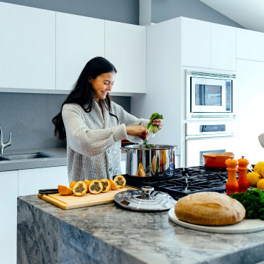 An image captures a young woman in the midst of meal preparation, surrounded by an array of fresh ingredients on the countertop of a modern home kitchen. Her focus and care in handling the food items reflect a passion for cooking and a commitment to healthy living.