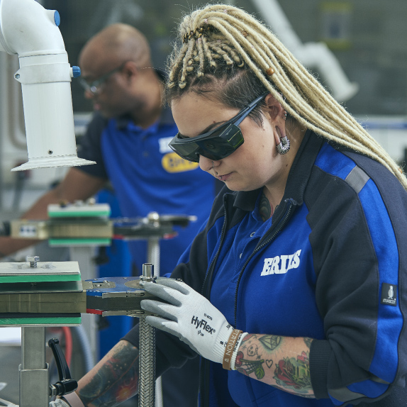 An image that celebrates diversity and inclusion in the workplace, showcasing a young woman with dreadlocks and tattoos, actively engaged in operating a high-tech machine in a factory setting. She is equipped with gloves and protective glasses, prioritizing safety while demonstrating her expertise. Dressed in a blue ERIKS jersey, her presence in this environment challenges traditional stereotypes and highlights the importance of embracing individuality and talent in all forms. The photograph captures a moment of skilled work, underlining the value of diversity and inclusion within the industry.