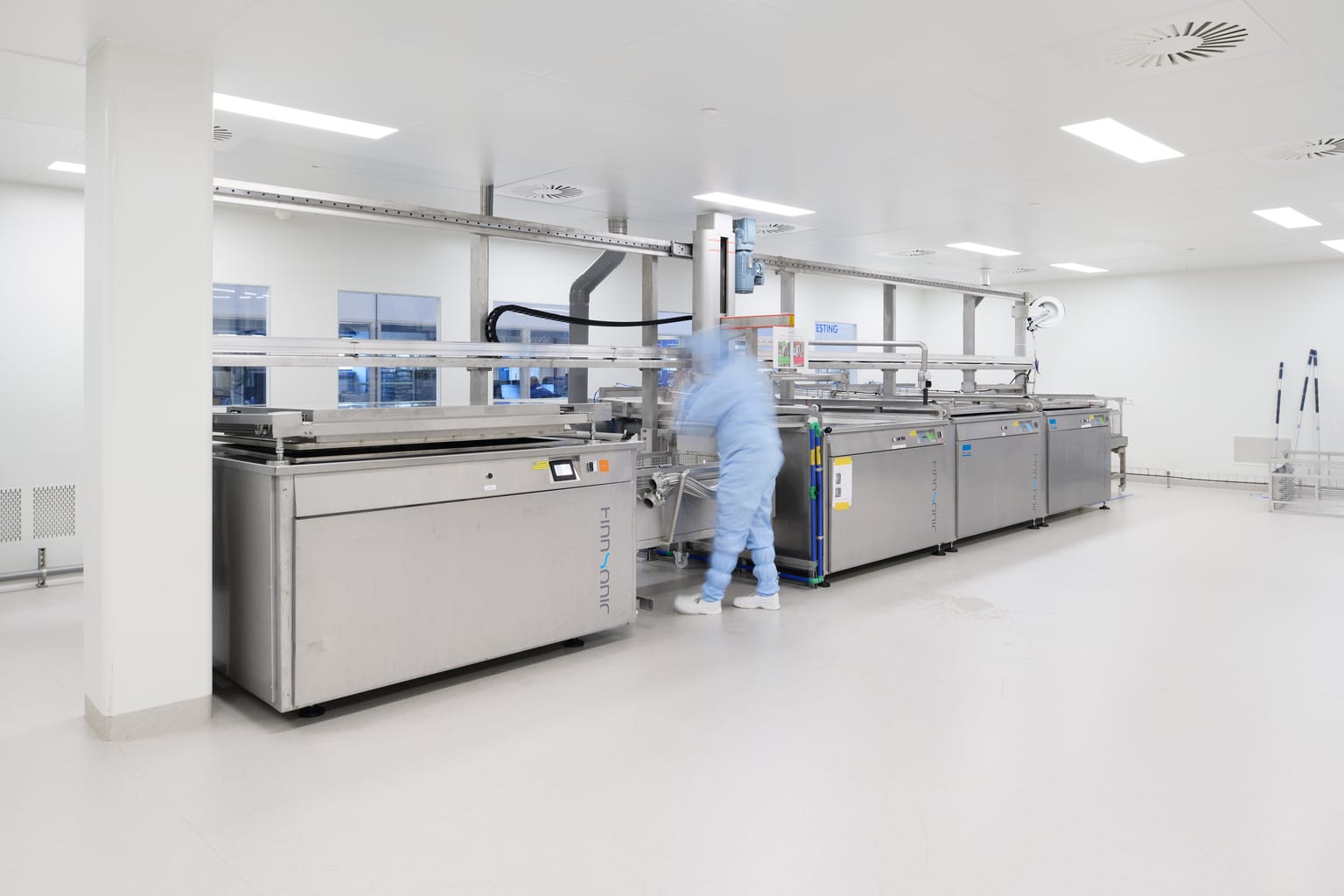 A person in blue cleanroom clothing stands beside large stainless steel manufacturing equipment in a bright, sterile production room. A person in blue cleanroom clothing stands beside large stainless steel manufacturing equipment in a bright, sterile production room.