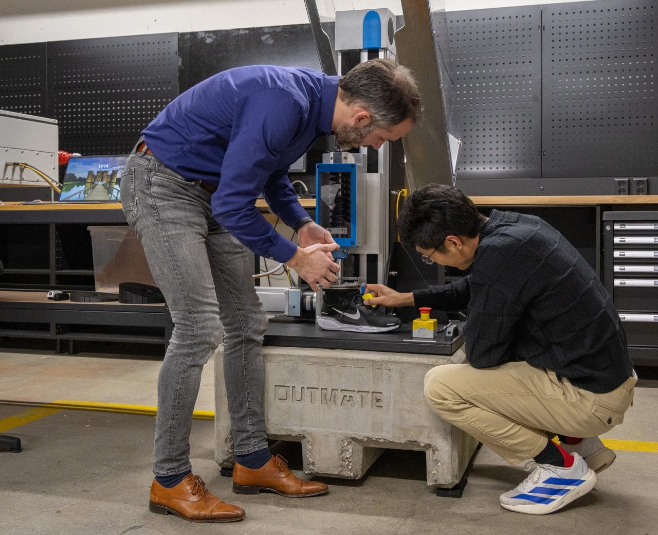 Two people test a shoe on a mechanical testing machine in a lab. One adjusts the device while the other operates the controls, with equipment and a workbench in the background. Two people test a shoe on a mechanical testing machine in a lab. One adjusts the device while the other operates the controls, with equipment and a workbench in the background.